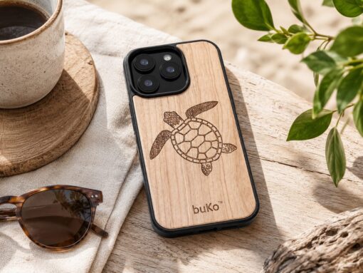 SEO file name: `buko-turtle-wood-phone-case-lifestyle-image.png` Alt tag: `BuKo wooden phone case with engraved turtle design on a natural beach-inspired table setting with sunglasses, coffee and greenery`