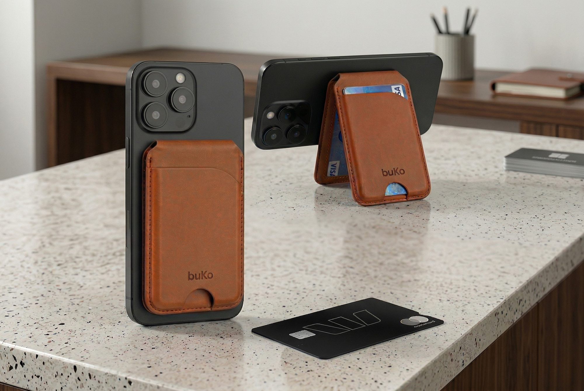 Black iPhones displayed with tan leather buKo MagSafe wallet stands on a stone countertop, with cards partially visible and a black bank card in the foreground.