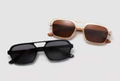 Two pairs of aviator-style sunglasses in black and transparent canary yellow frames with tinted lenses on a light grey background.